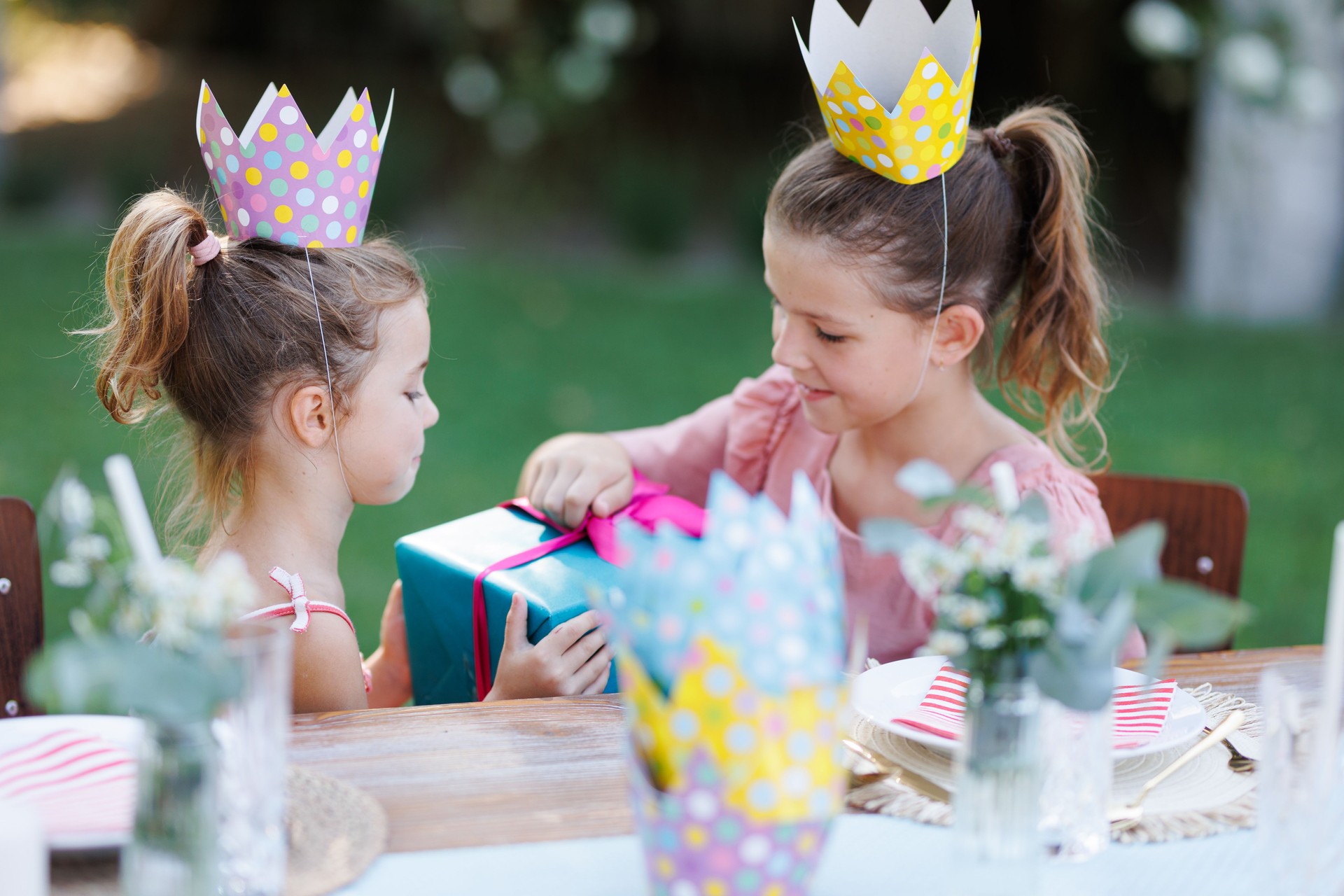 Piccoli amici alla festa di compleanno all'aperto in giardino. Ragazze carine con corone di carta che aprono il regalo di compleanno. Festa di compleanno in giardino per bambini.
